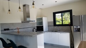 A modern kitchen with a central island, bar stools, a built-in oven, and a window with a view of greenery. - Porto Vecchio villa with heated pool near San Ciprianu beach (Porto Vecchio)