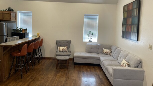 A modern living room with a grey sectional sofa, a small grey armchair, and a wooden dining table with bar stools. There are two windows with blinds, a kitchen area with a refrigerator and microwave, and a painting on the wall. - 4 bedrooms 3 bath, Tranquil Home, Master suite & Jet tub, Mins from Hot Springs (Thermopolis)