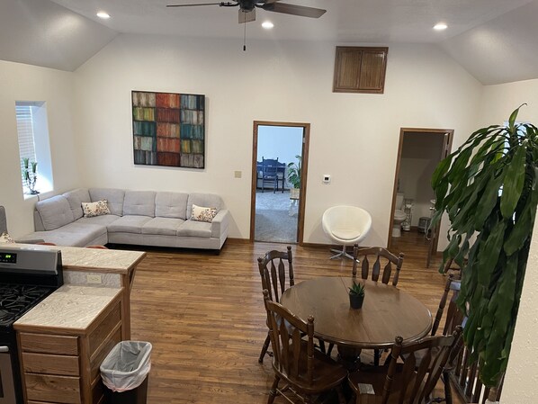 A modern living room with a grey sectional sofa, a dining table with wooden chairs, a white chair, a ceiling fan, and a large abstract painting.