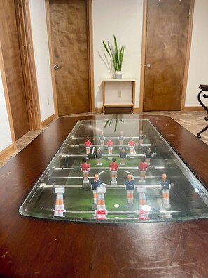 Une table de baby-foot dans une pièce dont le sol est en bois et où se trouve une plante sur une étagère.