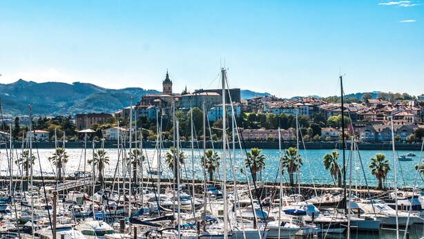 A marina with numerous boats docked, a prominent church tower in the background, and a coastal town with buildings and hills.