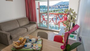 Ein modernes Wohnzimmer mit Sofa, Esstisch mit Frühstücksutensilien, einem Balkon mit roten Stühlen und Blick auf eine Berglandschaft.