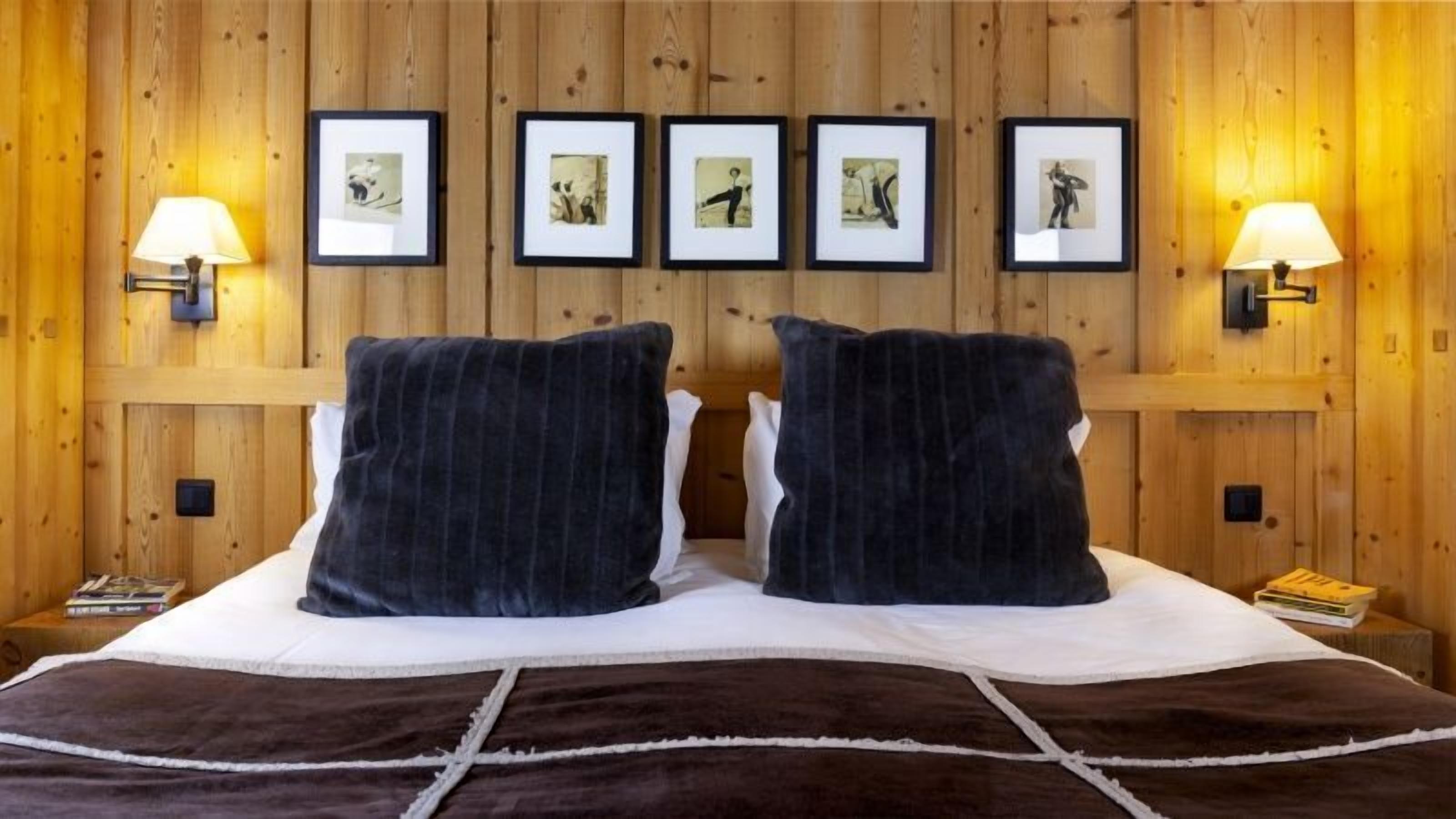 A hotel room with a wooden paneled wall, a bed with dark bedding, two bedside lamps, and framed pictures on the wall.