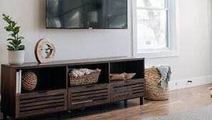 A modern living room with a large flat-screen TV mounted on the wall, a wooden console table with baskets, a potted plant, and a window providing natural light. - King beds, central location, entire house. Perfect for family gathering! (Saint Paul)