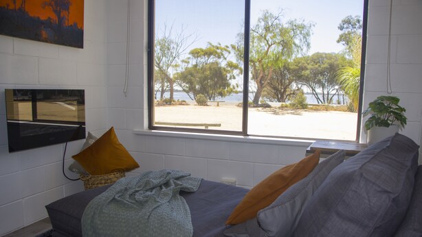 A room with a large window offering a view of a beach and trees. There is a gray sofa with a blanket and pillows, a small table, and a plant.