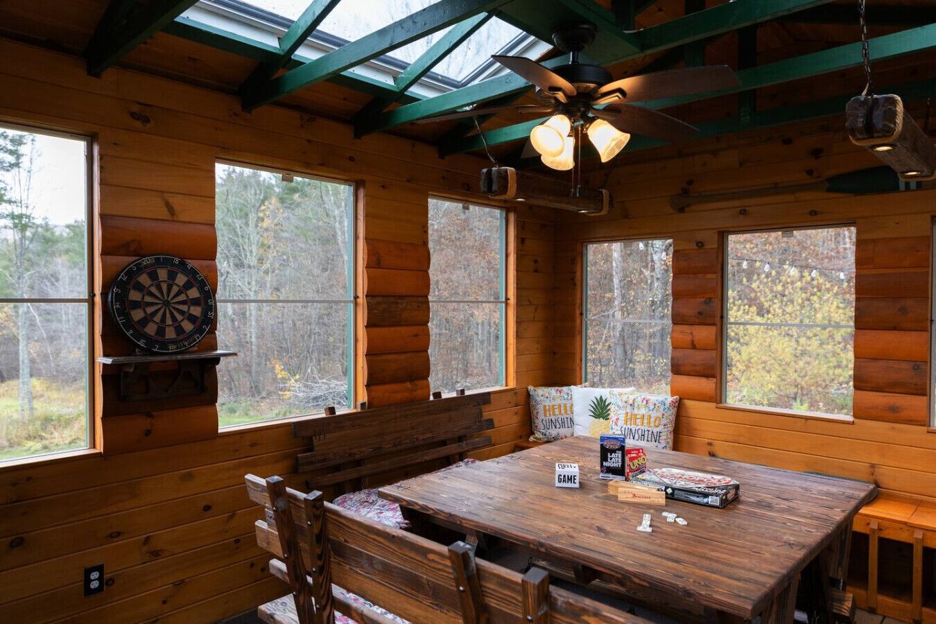 Interior de una cabaña de madera con una mesa de comedor, sillas, un dardo y un ventilador de techo.