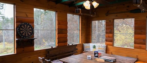 Interior de una cabaña de madera con una mesa de comedor, sillas, un dardo y un ventilador de techo.