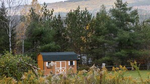 Eine kleine Holzhütte mit dunklem Dach, umgeben von dichten Bäumen und einer hügeligen Landschaft im Hintergrund.