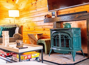 A cozy room with a wooden interior, a green stove, a television, and a table with board games. - Luxurious Mt Retreat Sauna Hot Tub Fire Pit & Park (Windham)