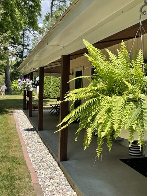 A covered patio area with a hanging fern plant, a concrete pathway, and a seating area with outdoor furniture. - Enjoy the beautiful views of the Muskegon River near The Hardy Dam/ Dragon Trail (Newaygo)