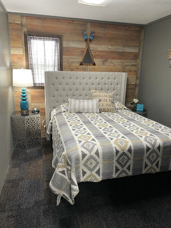 A bedroom with a tufted headboard, patterned bedding, a decorative wall, a window with curtains, a side table with a lamp, and a small sign with a motivational quote.