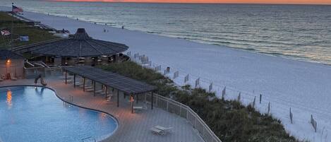 A beachside resort with a swimming pool, lounge chairs, and a view of the ocean at sunset.