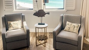 Una habitación con dos sillones, una mesita, un ventilador de techo y vistas al océano a través de ventanas grandes con contraventanas.