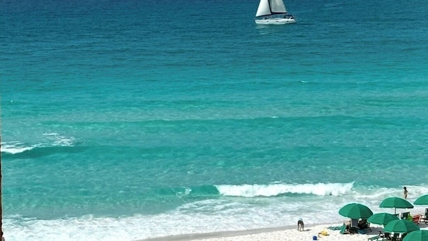 Una playa con agua turquesa cristalina, un velero y sombrillas.