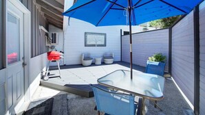 Eine Terrasse mit Tisch und Stühlen, einem blauen Sonnenschirm, einem Grill und einem kleinen Gartenbereich.