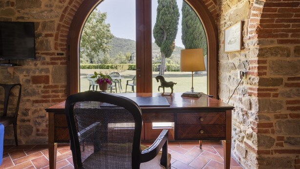 Quarto com parede de pedra, uma escrivaninha de madeira, uma cadeira, televisão fixada na parede e uma grande janela em arco com vista para um jardim.