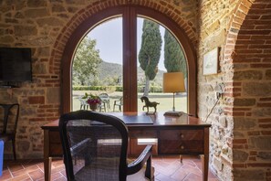 A room with a stone wall, a wooden desk, a chair, a TV mounted on the wall, and a large arched window overlooking a garden.