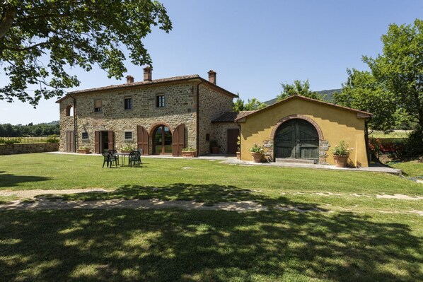 A stone house with a large lawn, a small attached building with a door, and a table with chairs.