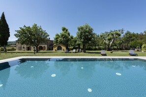 A clear swimming pool with lounge chairs, a stone building, and trees in the background.