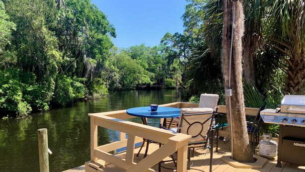 Une terrasse en bois avec une table et des chaises, donnant sur une rivière et entourée d’une végétation luxuriante.