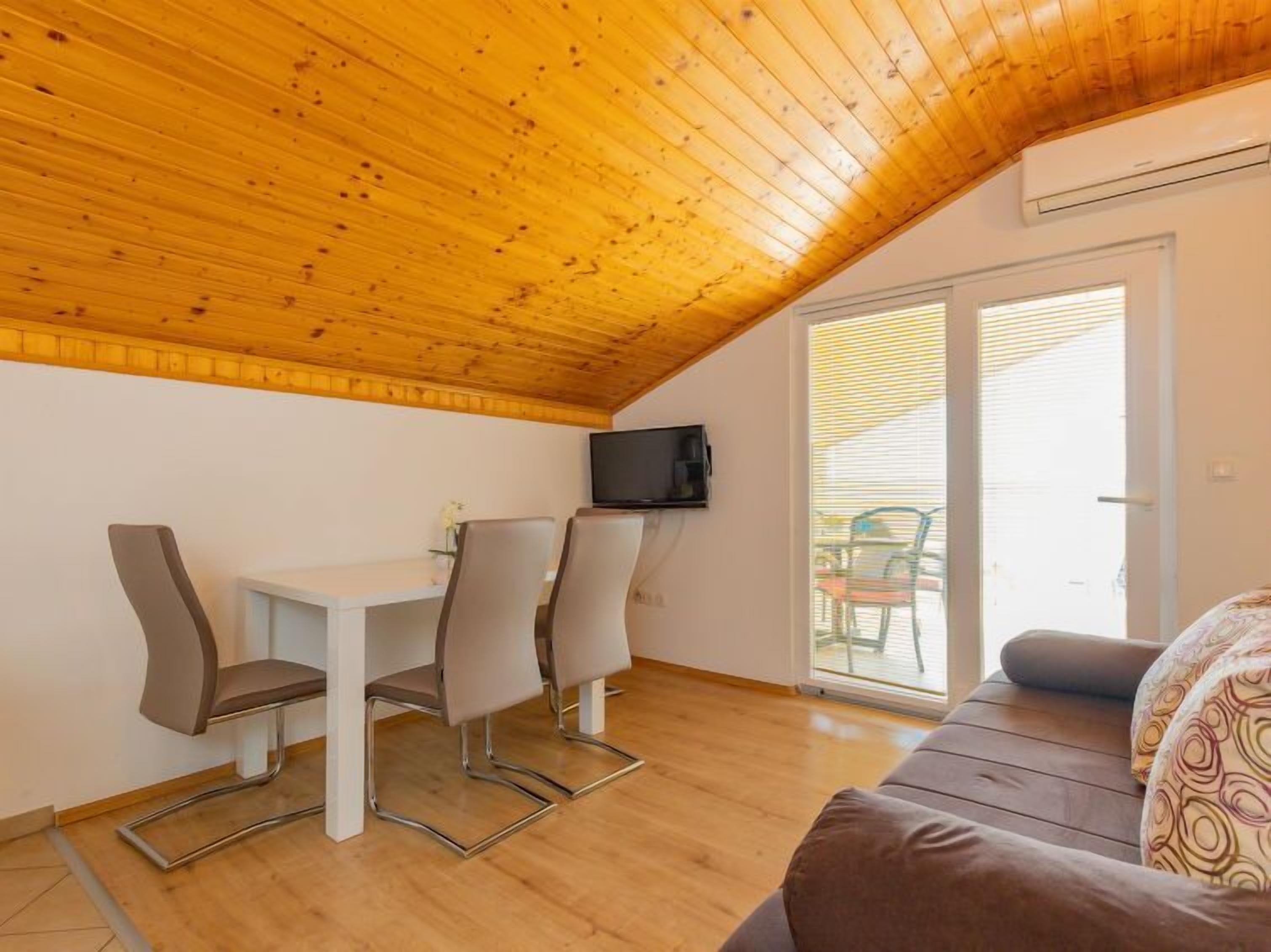 A cozy room with a wooden ceiling, a dining table with chairs, a sofa, and a TV mounted on the wall.