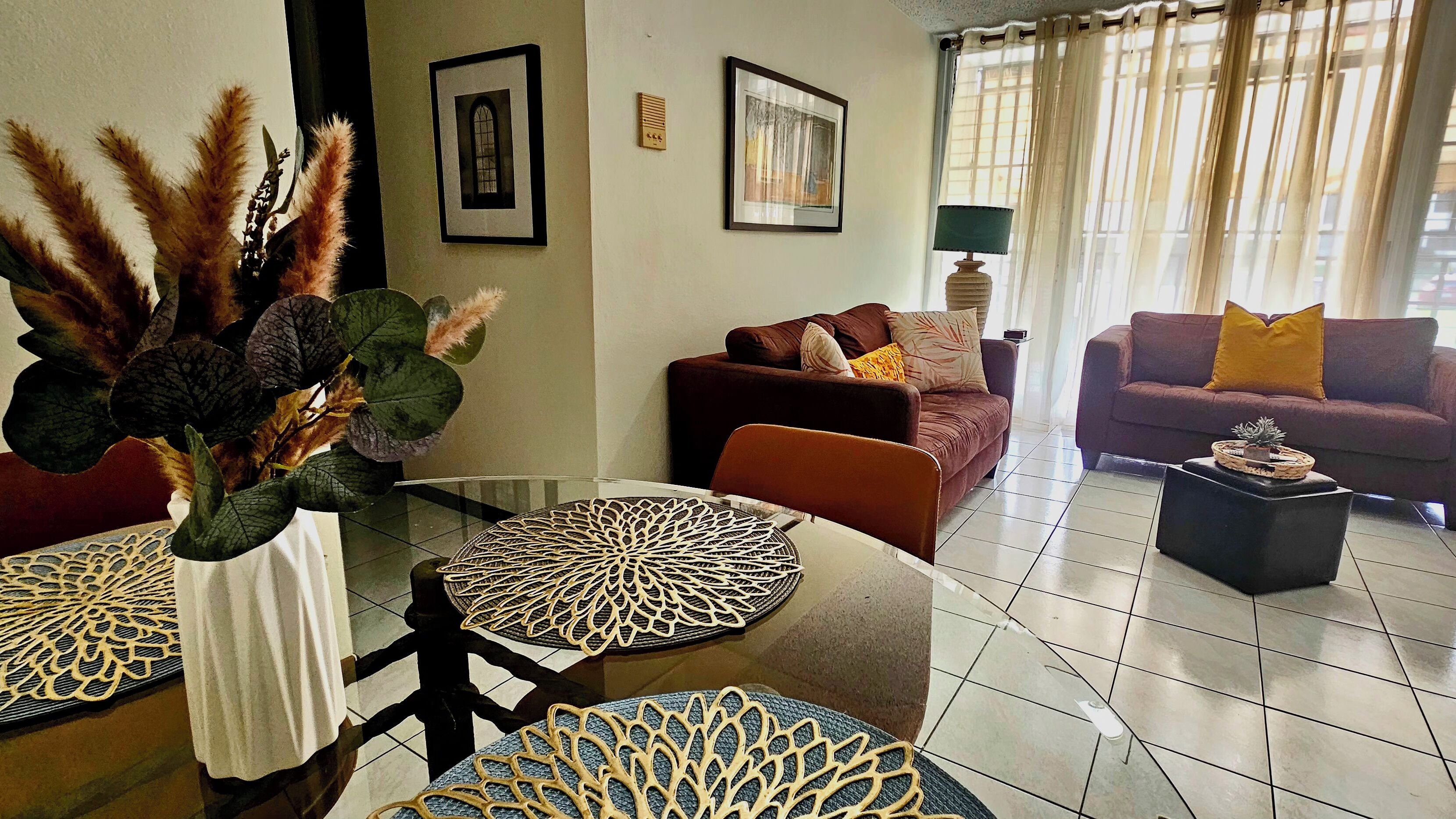 A living room with two sofas, a coffee table, a glass table with a decorative centerpiece, and a large window with curtains.