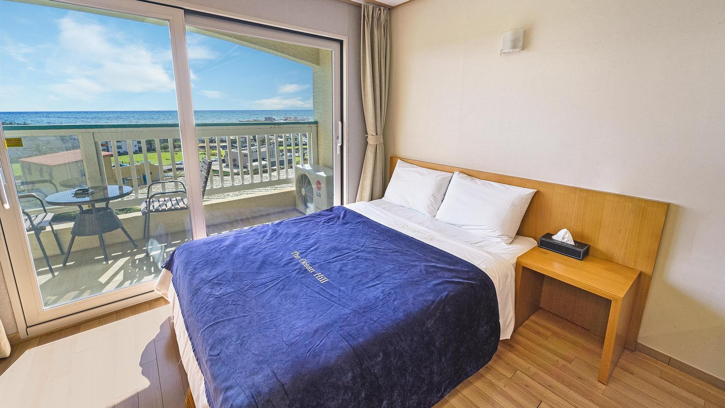 A hotel room with a large balcony, a bed with a blue cover, and a wooden headboard.