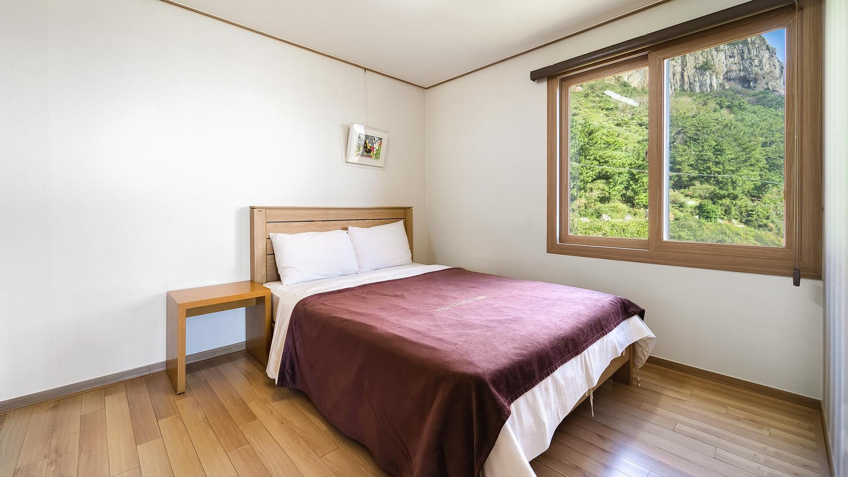 A bedroom with a wooden bed, a nightstand, a window with a view of greenery, and a framed picture on the wall.