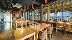 Um restaurante com mesas e cadeiras de madeira, grandes janelas com vista e luminárias pendentes.