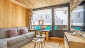 Una sala de estar acogedora con un sofá gris, una pequeña mesa de madera con una revista, y vistas a un balcón con sillas coloridas.