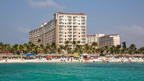 Een groot hotel met meerdere verdiepingen en balkons, gelegen aan een strand met palmbomen en een menigte mensen.
