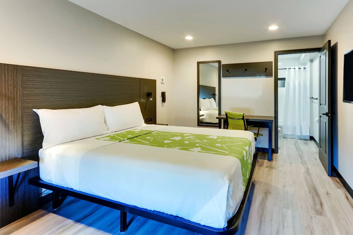 Habitación de hotel moderna con una cama grande, un escritorio, un espejo y un baño visible a través de una puerta abierta.