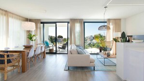 Una sala de estar moderna con sofá, mesa de comedor y sillas. Hay un ventanal con vistas a edificios y vegetación.