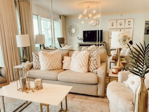 A living room with a beige sofa, a coffee table, and a TV. There are decorative pillows, a chandelier, and wall art. - Beach Haven •Amazing View•Lagoon Pool •Luxury Condo (Destin)