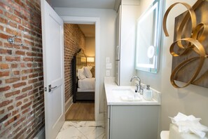 Une salle de bain avec un lavabo blanc, un miroir et des toilettes. Une porte donne sur une chambre à coucher qui contient un lit et une lampe. La salle de bain est dotée d’un sol en marbre et d’un mur en briques.