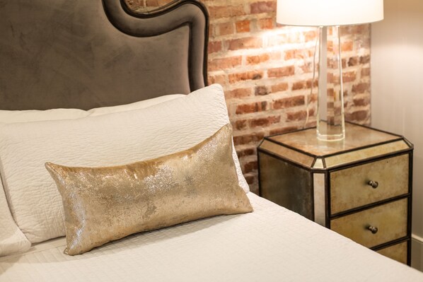 Un lit bien fait, agrémenté d’un coussin blanc texturé et d’un coussin décoratif doré. Une lampe moderne à abat-jour transparent est posée sur une petite table d’appoint à côté du lit. La pièce est dotée d’un mur en briques et d’une tête de lit sombre.