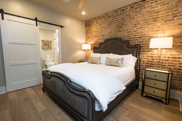 Un dormitorio con pared de ladrillo, una cama con ropa de cama blanca, dos mesitas de noche con lámparas y una puerta corredera.