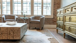 Ein Zimmer mit einem großen Fenster, einem Holztisch, zwei Sesseln, einem weißen Fellteppich und einer Kommode im Vintage-Stil.