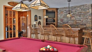 Ein Billardtisch mit roter Spielbahn, Holzstühle und ein Barbereich mit einer Wand aus Stein im Hintergrund.