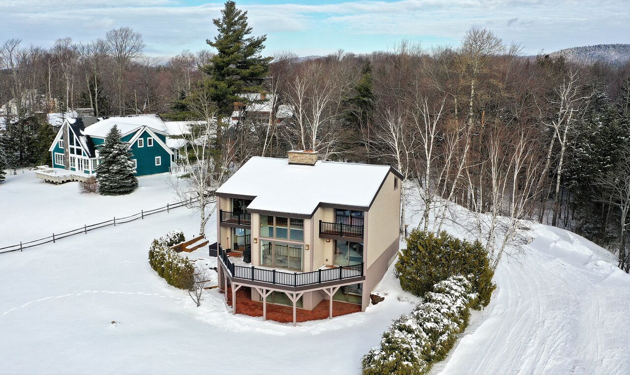 A large, two-story house with a balcony, surrounded by snow-covered landscape and trees.