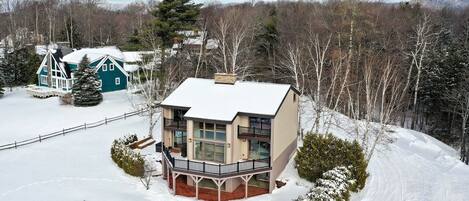 A large, two-story house with a balcony, surrounded by snow-covered landscape and trees.