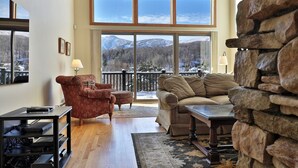 Ein Wohnzimmer mit großem Fenster und Blick auf die Berge, einer Steinwand, einer Couch, einem roten Sessel und einem TV-Schrank.