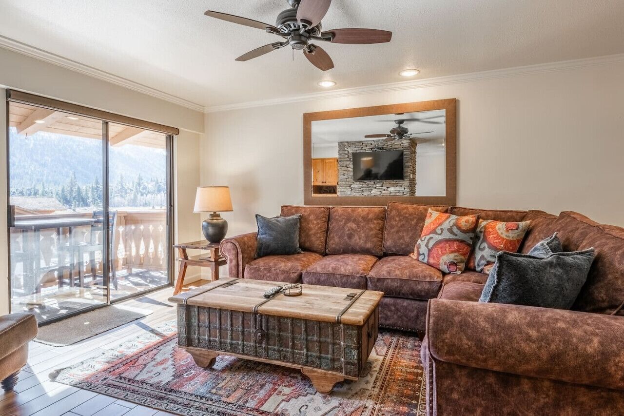A living room with a brown leather sofa, a wooden coffee table, a floor lamp, and a large window with a view of mountains.