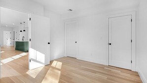 Un hall spacieux et bien éclairé, doté d’un plancher en bois et de murs blancs, qui mène à plusieurs portes et donne sur un espace cuisine visible.
