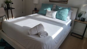 Un lit bien fait, avec du linge de lit blanc et une serviette pliée, une table de chevet avec une lampe, une petite plante en pot et un tableau accroché au mur.