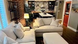 A modern living room with a sectional sofa, a dining area with a kitchen in the background, and a brick wall. - Fully Remodeled Home In Boulder County (Superior)