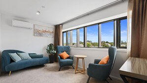 A modern living room with a blue sofa, two armchairs, a small round table, and a large window offering a view of the outdoors. - Golden Hotel (Christchurch)
