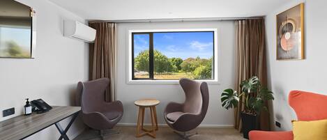 A modern hotel room with a desk, two chairs, a small table, a sofa, a potted plant, and a large window with a view of greenery.