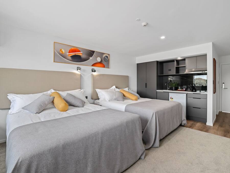 A modern hotel room with two beds, a kitchenette, and a large painting on the wall.