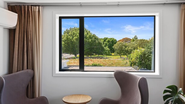 A room with a large window offering a view of a park and trees. There is a chair, a small table, and an air conditioning unit. - Golden Hotel (Christchurch)